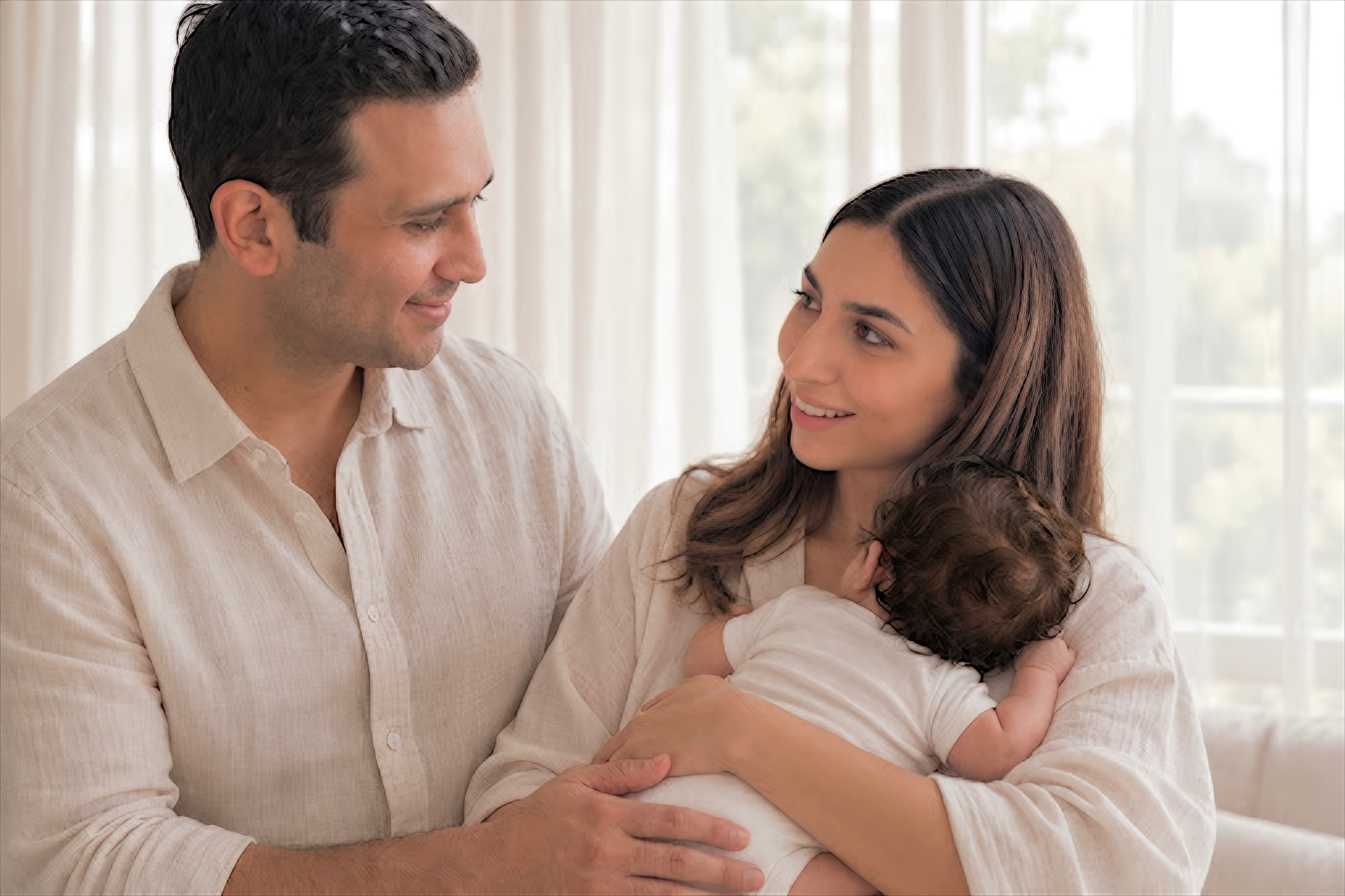 A smiling father and mother gazing at each other as the mother cradles their newborn by a bright sunlit window